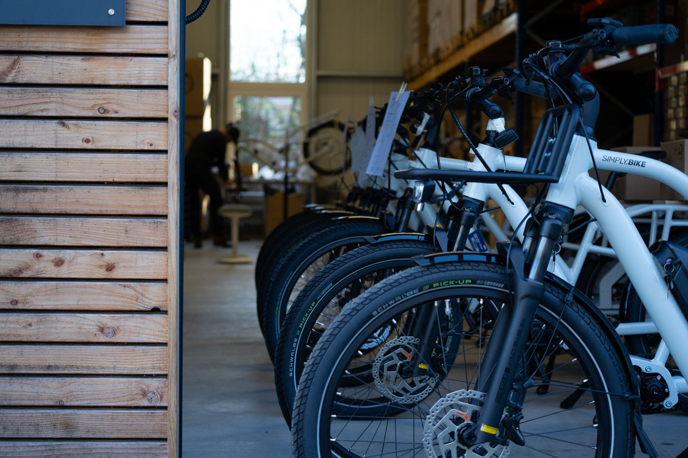 Reihe weißer Simply.Bike E-Lastenräder in der Werkstatt in Hamburg-Bergstedt, im Hintergrund ein Mechaniker bei der Reparatur.
