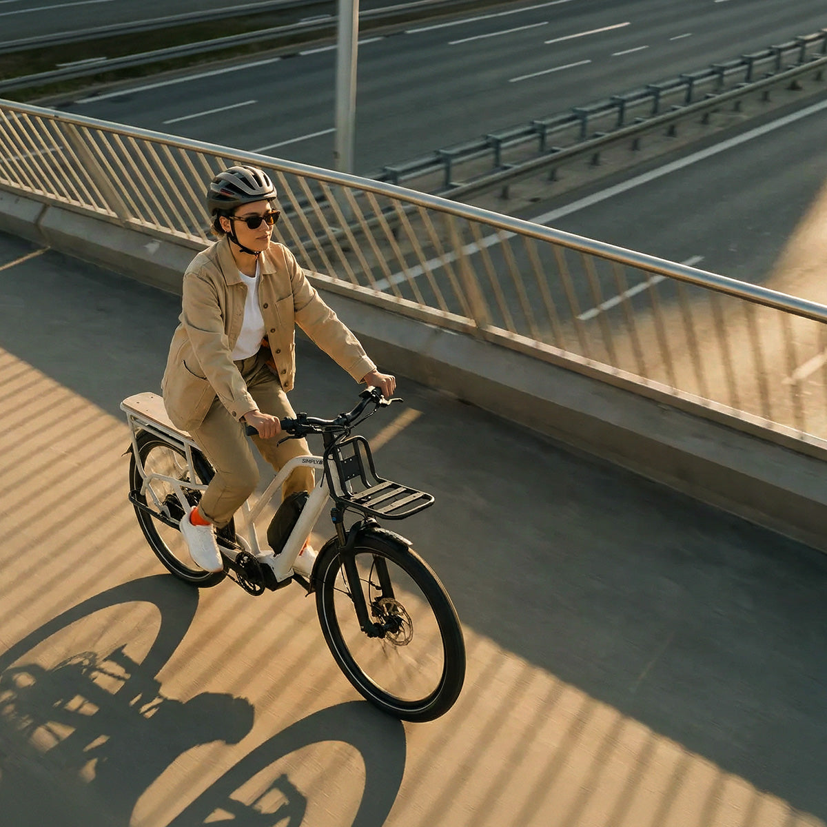 Eine Person fährt ein Simply.Lift EP6 Longtail E-Lastenrad mit Frontgepäckträger auf einem Radweg über einer mehrspurigen Straße.