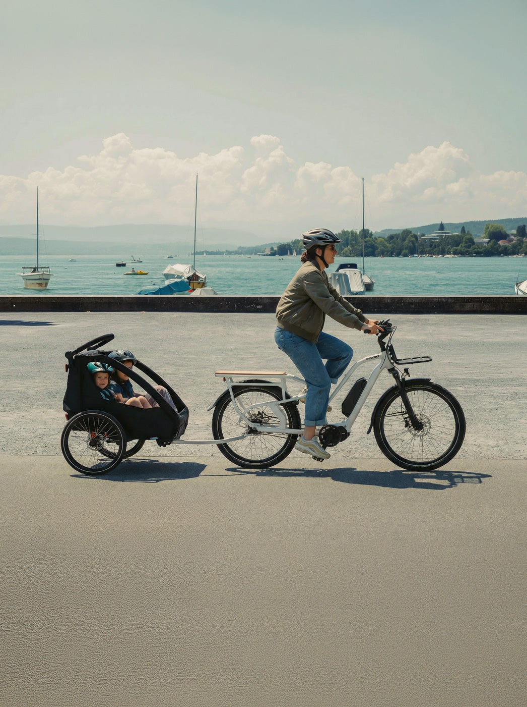 Eine Person fährt ein weißes Simply.Lift EP6 Longtail E-Lastenrad mit einem angekoppelten Kinderanhänger, in dem zwei Kinder sitzen, auf einer Uferpromenade am See.
