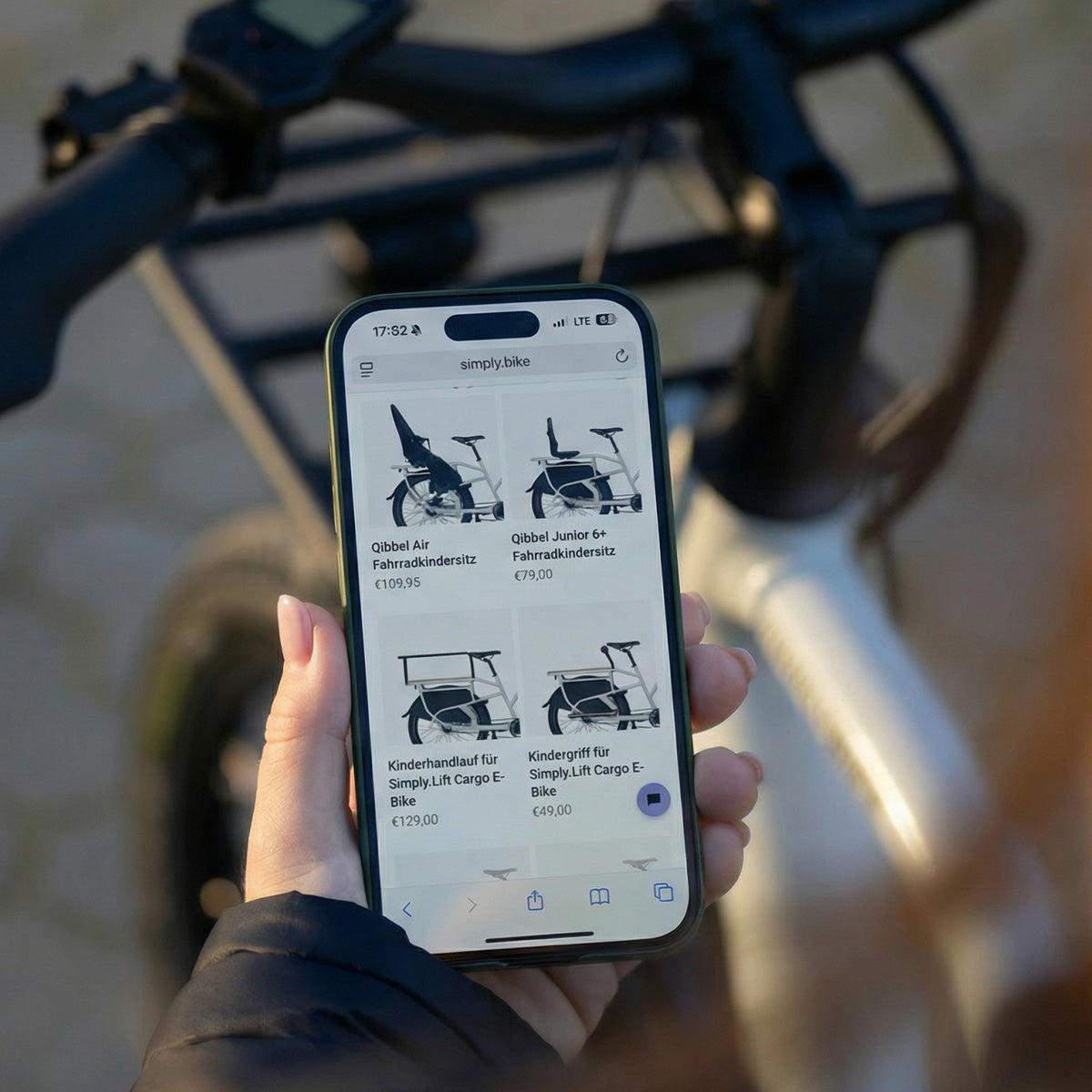 Smartphone mit dem Simply.Bike Online-Shop hält; auf dem Display ist Zubehör für das Simply.Lift E-Lastenrad wie Kindersitze und Kinderhandläufe zu sehen.
