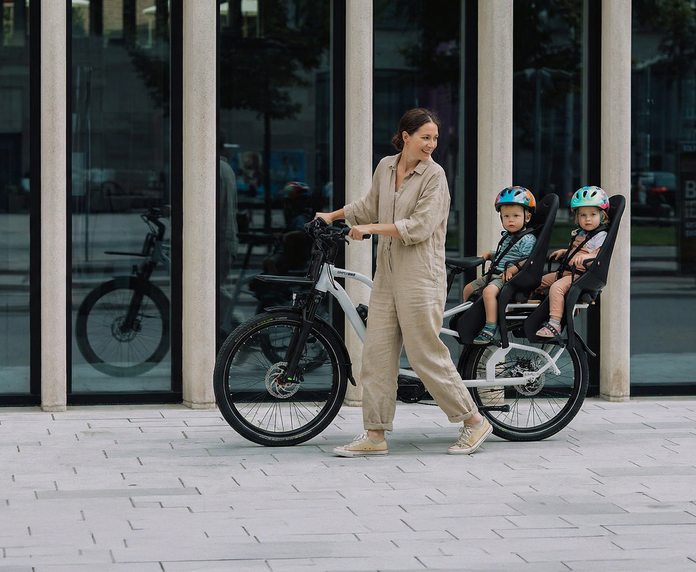 Eine Frau schiebt ein Simply.Lift EP6 Longtail E-Lastenrad mit zwei Kindern in hinteren Kindersitzen und einem Frontgepäckträger vor einer modernen Glasfassade.