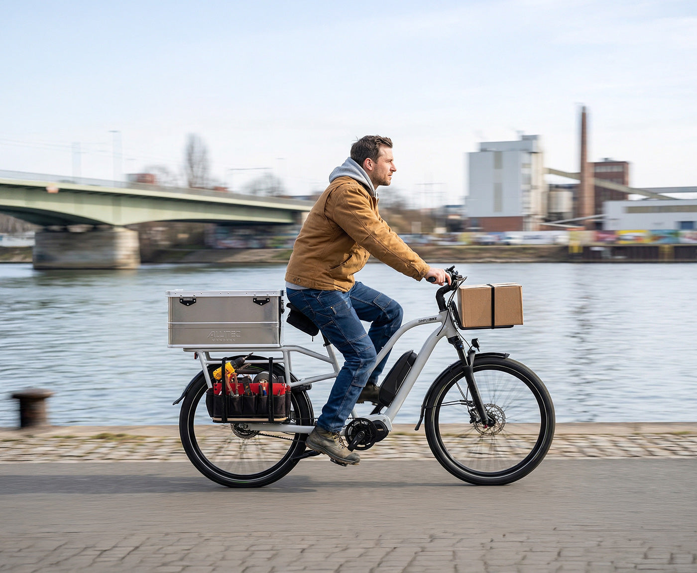 Ein Mann fährt ein Simply.Lift EP6 Longtail E-Lastenrad mit einer großen Transportbox aus Aluminium, seitlich montierten Werkzeugtaschen und einem Paket auf dem Frontgepäckträger entlang einer Uferpromenade.