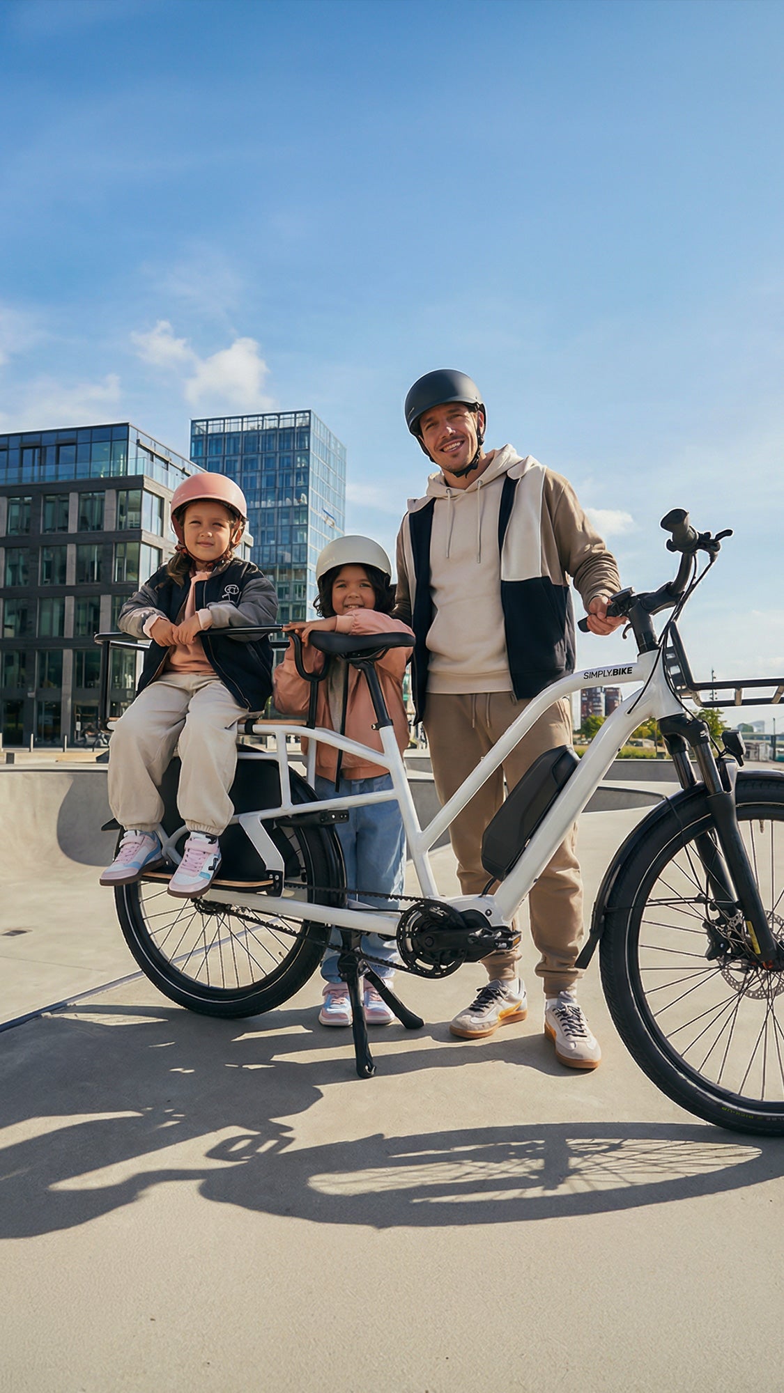 Ein Mann steht mit zwei Kindern an einem weißen Simply.Lift EP6 Longtail E-Lastenrad auf einer sonnigen Uferpromenade vor Glasgebäuden.