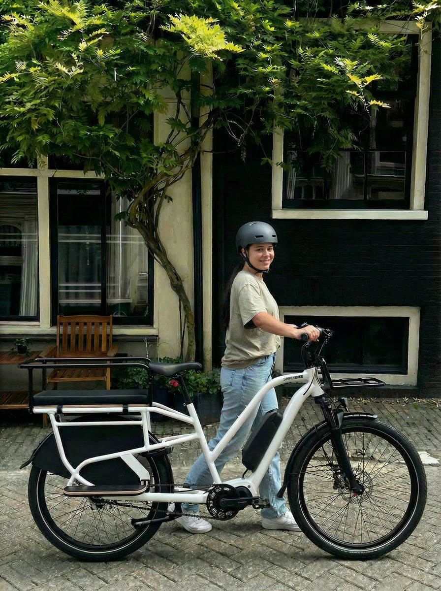 Lächelnde Frau mit Helm schiebt ein Simply.Lift EP6 Longtail E-Lastenrad mit montiertem Kinderhandlauf und Frontgepäckträger vor einem Haus mit Wandbegrünung.