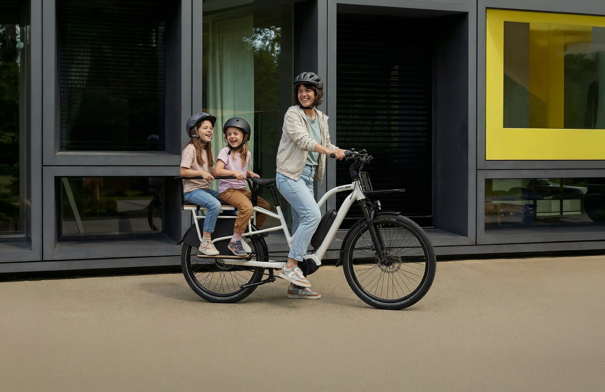 Zwei Kinder sitzen mit Helmen gesichert auf dem Gepäckträger eines weißen Simply.Lift EP6 Longtail E-Lastenrads mit Kinderhandlauf.