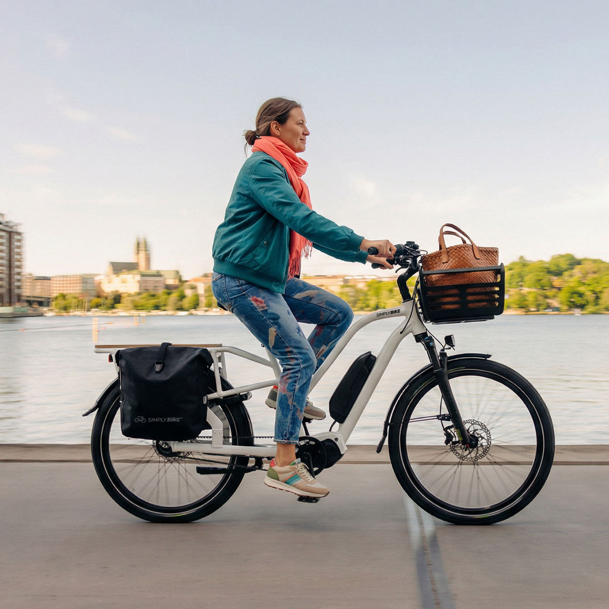 Frau fährt weißes Simply.Lift E-Lastenrad mit Thule Pack 'n Pedal Fahrradkorb am Wasser