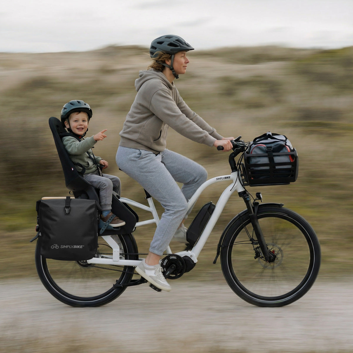 Frau fährt ein Simply.Lift EP6 Longtail E-Lastenrad mit Kind im Rücksitz, seitlicher Packtasche und Frontkorb auf einem unbefestigten Weg.