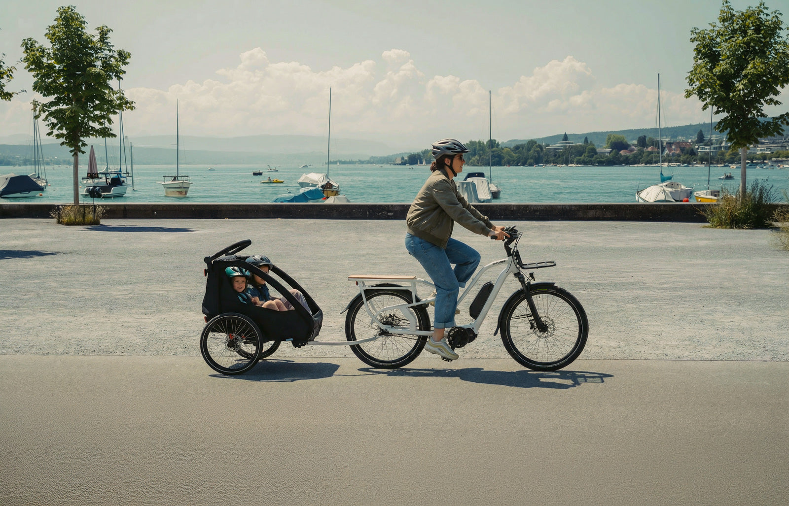 Simply.Lift EP6 Longtail E-Lastenrad mit angekoppeltem Kinderanhänger während einer Fahrt auf einer Uferpromenade am See.