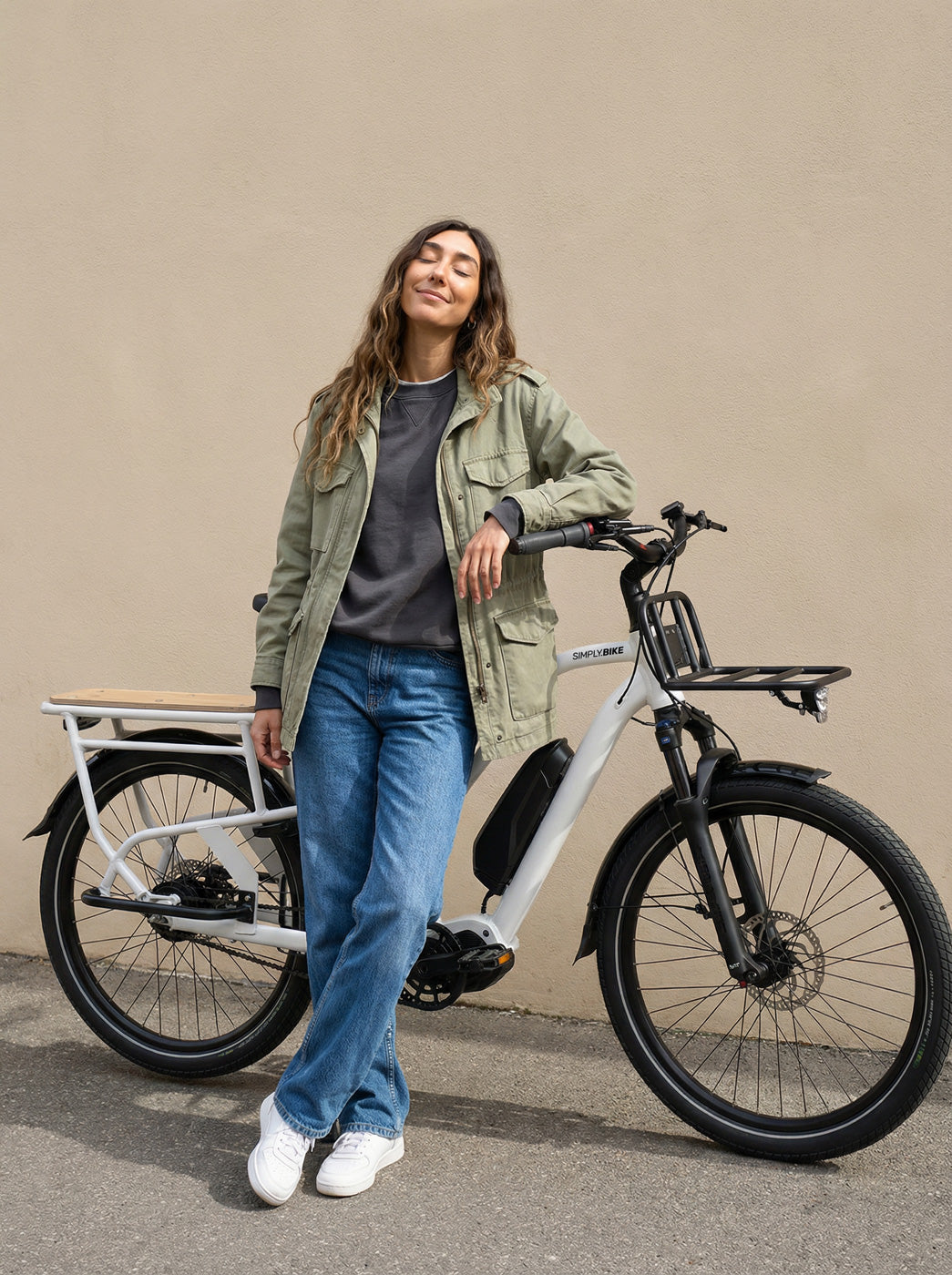 Frau lehnt entspannt an einem Simply.Lift EP6 Longtail E-Lastenrad mit Frontgepäckträger und Mittelmotor vor einer hellen Wand.