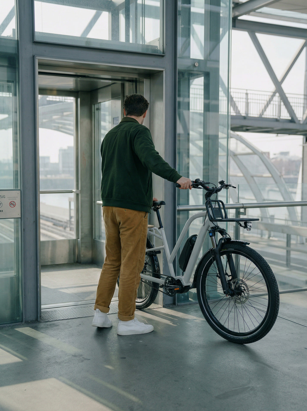 Mann schiebt ein Simply.Lift EP6 Longtail E-Lastenrad mit Frontgepäckträger in einen gläsernen Aufzug zur Mitnahme im urbanen Raum.