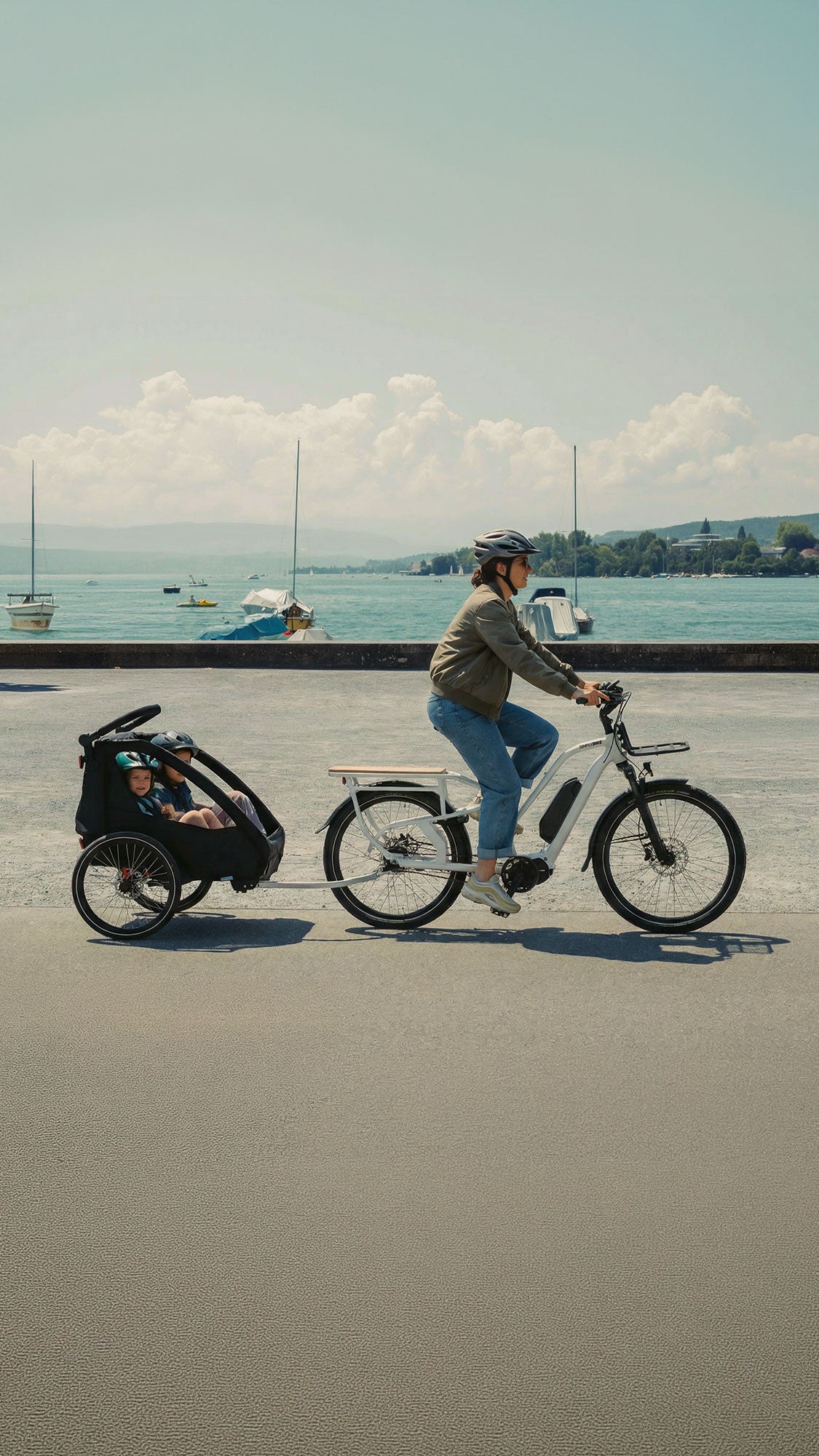 Simply.Lift EP6 Longtail E-Lastenrad mit angekoppeltem Kinderanhänger während einer Fahrt auf einer Uferpromenade am See.