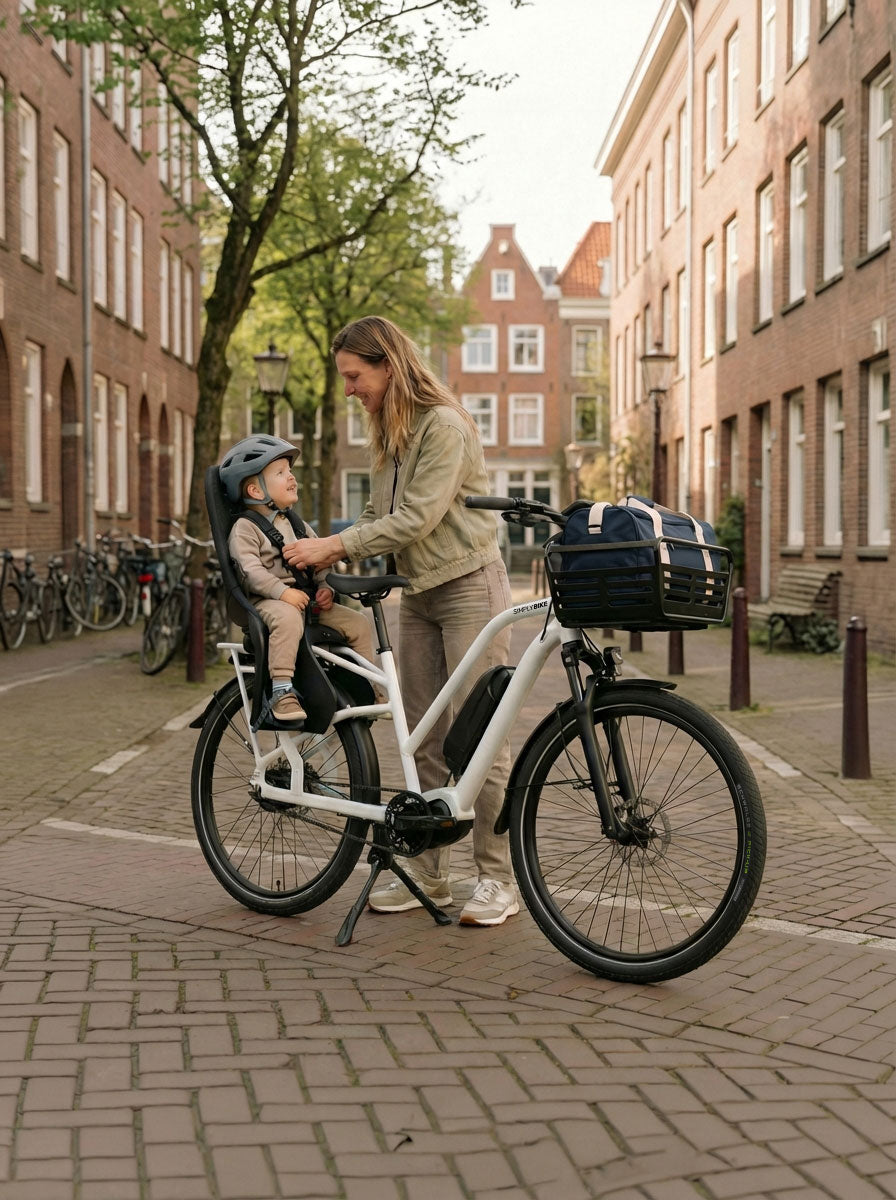 Mutter schnallt Kleinkind im Kindersitz eines Simply.Lift EP6 Longtail E-Lastenrads fest; am Frontgepäckträger ist ein Korb mit Transporttasche montiert.