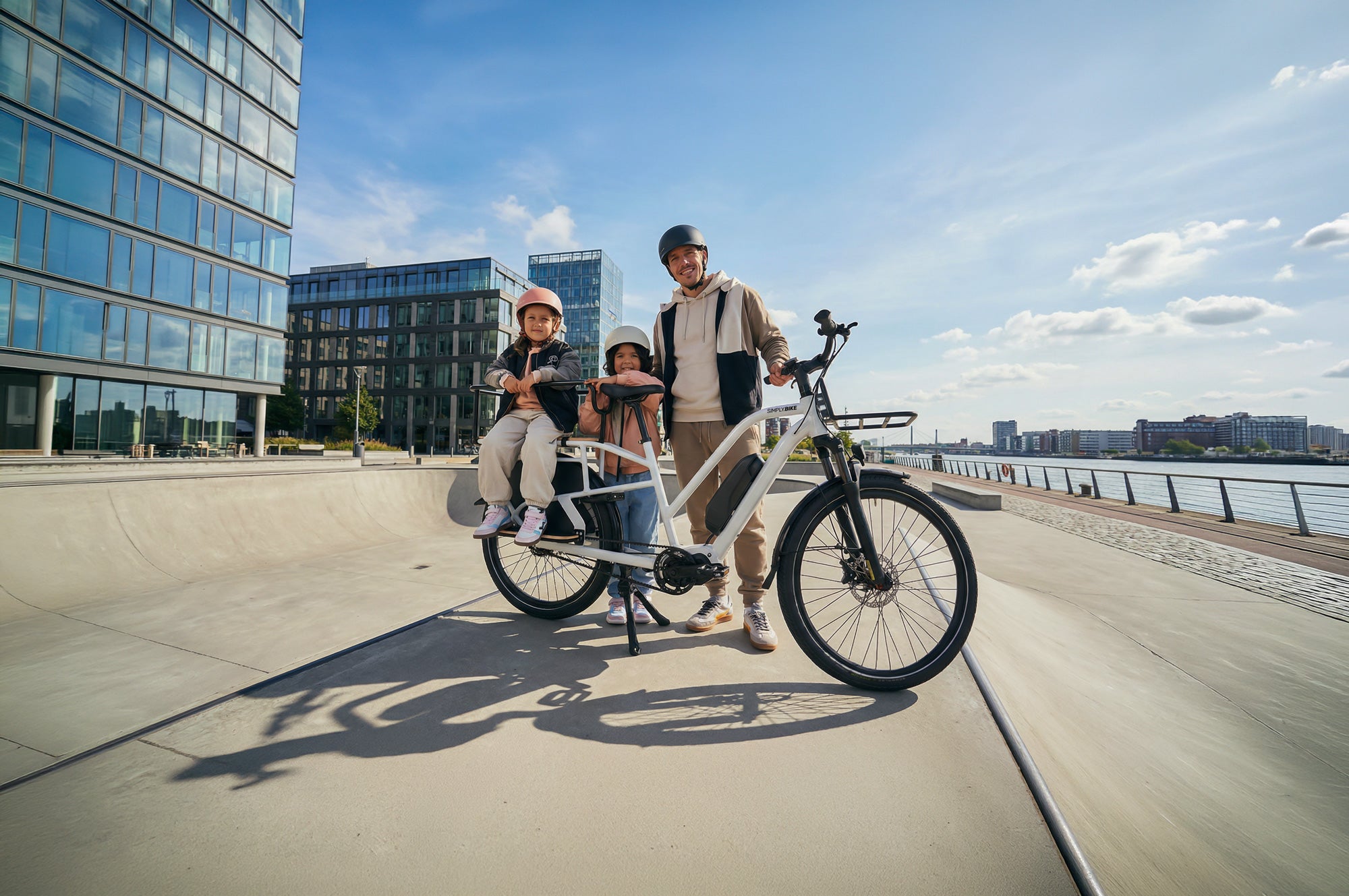 Ein Mann steht mit zwei Kindern an einem weißen Simply.Lift EP6 Longtail E-Lastenrad auf einer sonnigen Uferpromenade vor Glasgebäuden.