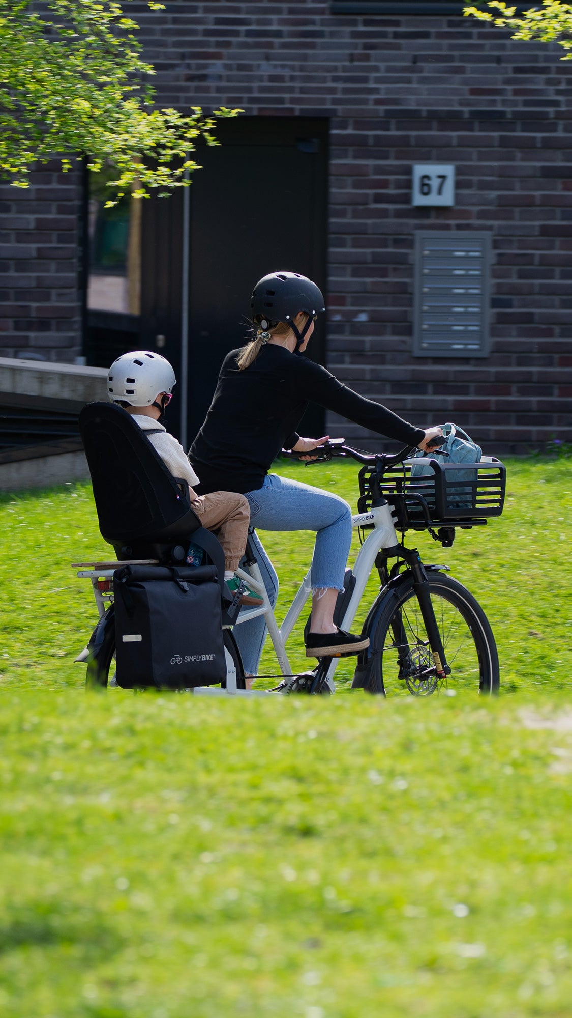 Frau fährt mit Kind im Kindersitz auf dem weißen Simply.Lift EP6 E-Lastenrad, mit Packtasche und Korb.