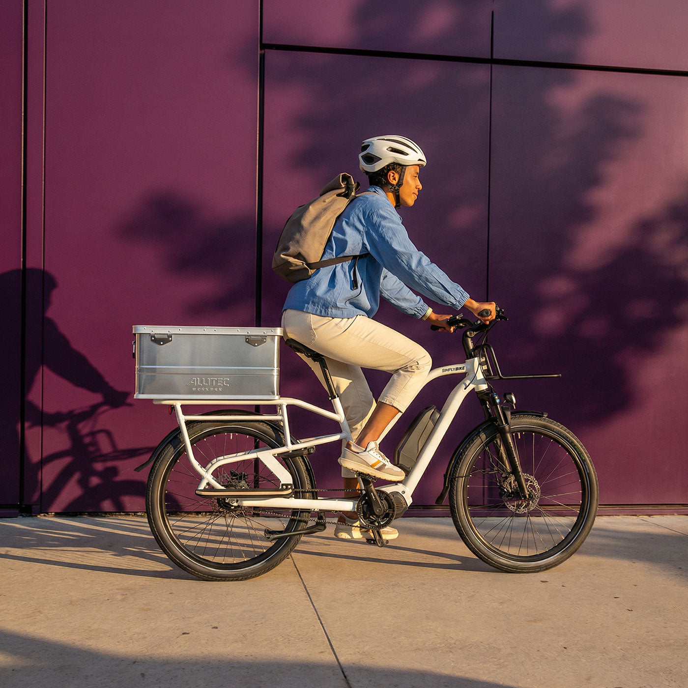 Frau fährt das weiße Simply.Lift EP6 Longtail E-Lastenrad mit Alukiste auf dem Gepäckträger vor einer Wand.
