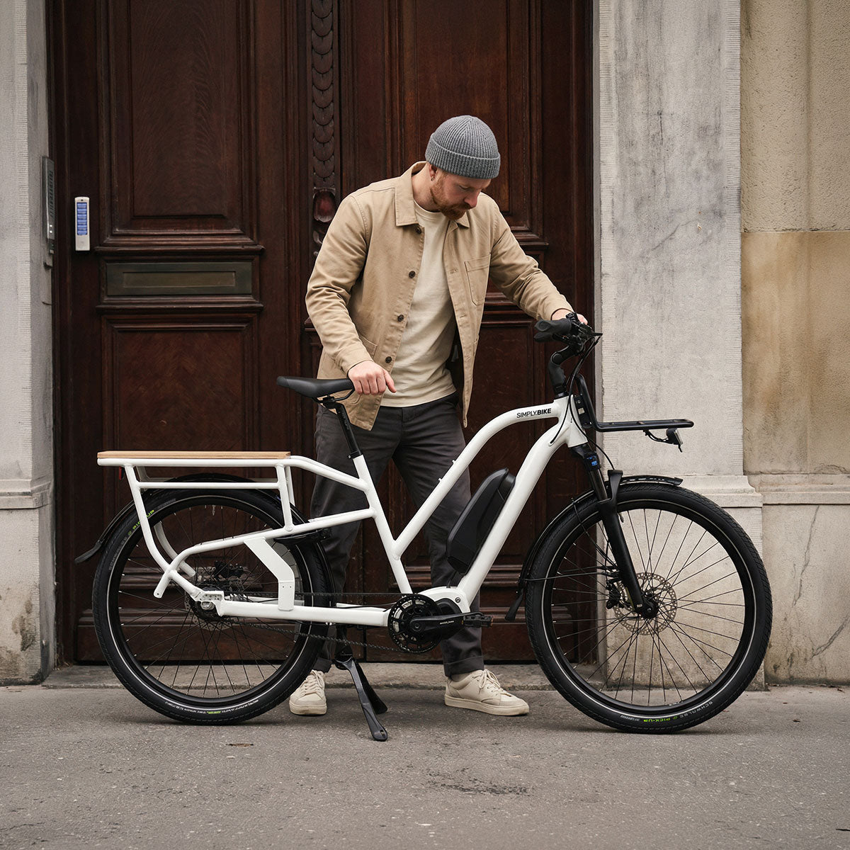 Mann mit Mütze schiebt das weiße Simply.Lift EP6 E-Lastenrad vor einer Altbautür in der Stadt.